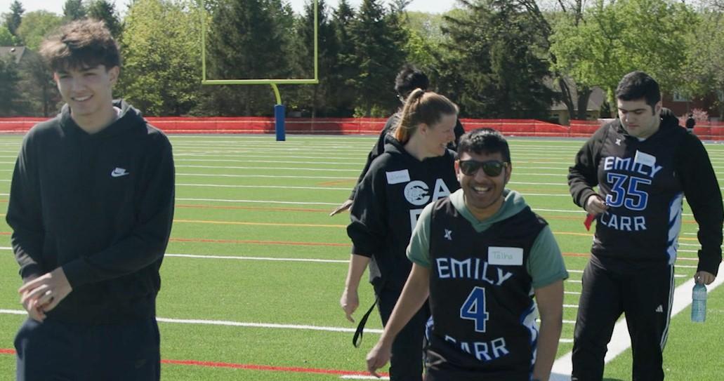 Four students stand on turf field smiling, walking towards the camera wearing blue sport shirts